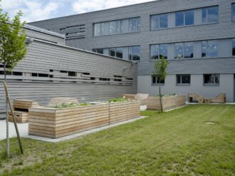 Ein moderner Schulgarten im Innenhof eines Gebäudes mit grauer Holzfassade und großen Fenstern, die mit weißen Vogelstickern dekoriert sind. Mehrere rechteckige Hochbeete aus hellem Holz sind mit jungen Pflanzen bepflanzt. Dazwischen stehen ebenfalls aus Holz gefertigte Sitzbänke mit Rückenlehnen, die ein geometrisches Design haben. Zwei frisch gepflanzte Bäume wachsen im Rasenbereich, der den Garten umgibt. Die Umgebung wirkt gepflegt und freundlich, ideal für Outdoor-Lernaktivitäten und Pausen im Freien.