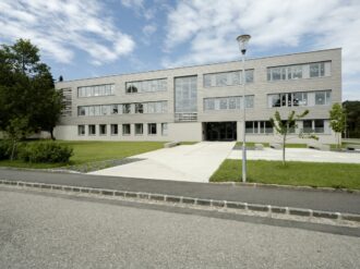 Vorderansicht eines modernen, dreistöckigen Gebäudes mit grauer Holzfassade und großen Fenstern, vor dem sich eine gepflasterte Einfahrt mit einem Gehweg befindet. Eine Straßenlaterne und mehrere junge Bäume stehen in der gepflegten Grünanlage vor dem Gebäude, während der Himmel teils bewölkt ist.