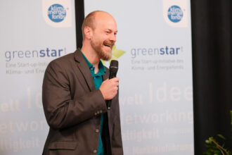 Ein Mann mit Glatze und Bart hält ein Mikrofon und spricht lächelnd vor einem „greenstart“-Banner, einer Start-up-Initiative für Klima- und Energielösungen. Er trägt ein dunkelgraues Sakko über einem türkisfarbenen Hemd und wirkt engagiert, während er auf der Bühne präsentiert. Der Hintergrund ist leicht verschwommen, mit schwarzen Vorhängen und einigen Pflanzen.