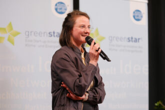 Eine junge Frau mit Brille und dunkler Jacke spricht auf einer Bühne in ein Mikrofon, während sie lächelt. Hinter ihr sind „greenstart“-Banner zu sehen, die auf eine Start-up-Initiative im Bereich Klima- und Energielösungen hinweisen. Ihre Körpersprache wirkt selbstbewusst und engagiert.