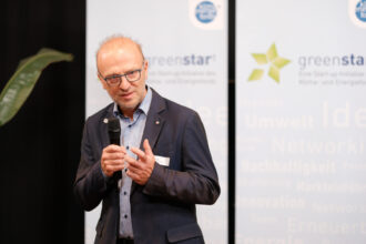 Ein Mann in einem dunkelblauen Sakko mit Brille hält ein Mikrofon und spricht auf einer Veranstaltung. Im Hintergrund sind Banner mit dem Logo von „greenstar“ und „klima+energie fonds“ zu sehen, die auf eine Konferenz oder ein Networking-Event im Bereich Klima und Energie hinweisen. Seine Gestik und Mimik lassen auf eine engagierte Präsentation oder Diskussion schließen.