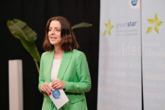 Eine Frau in einem grünen Blazer steht auf einer Bühne und spricht mit einem Lächeln ins Publikum. Sie trägt ein Headset-Mikrofon und hält eine Karte mit dem Logo von „Klima- und Energiefonds“ in der Hand. Im Hintergrund sind „greenstart“-Banner zu sehen, was auf eine Veranstaltung im Bereich Nachhaltigkeit und Innovation hinweist.