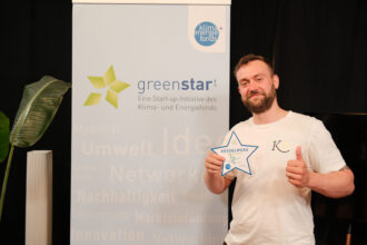 Ein Mann mit Bart und kurzem Haar steht vor einem „greenstart“-Banner und hält eine sternförmige Auszeichnung mit der Aufschrift „KESSELWERK Start-up TOP 10“ in der Hand. Er trägt ein weißes T-Shirt mit einem Logo auf der Brust und zeigt mit der anderen Hand einen Daumen hoch. Die Szene wirkt wie eine Preisverleihung im Bereich nachhaltiger Start-ups.