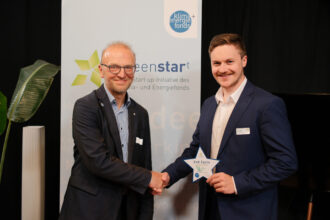 Zwei Männer stehen vor einem „greenstart“-Banner und schütteln sich die Hände. Der Mann auf der linken Seite trägt eine Brille und einen dunklen Anzug, während der Mann auf der rechten Seite einen dunkelblauen Anzug mit einem weißen Hemd trägt. Er hält eine sternförmige Auszeichnung mit der Aufschrift „2nd Cycle“ in der Hand und lächelt in die Kamera.