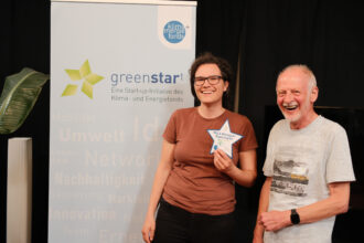 Eine junge Frau mit Brille und braunem T-Shirt steht lachend vor einem „greenstart“-Banner und hält eine sternförmige Auszeichnung mit der Aufschrift „MILA Mitmach-Supermarkt“ in der Hand. Neben ihr steht ein älterer Mann mit einem weißen gestreiften T-Shirt, der ebenfalls breit lacht. Beide wirken erfreut über die Ehrung.