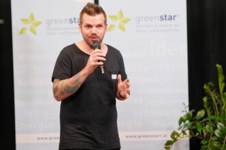 Ein Mann mit einem dunklen T-Shirt und tätowiertem Arm spricht in ein Mikrofon auf einer Veranstaltung mit einem „greenstart“-Banner im Hintergrund. Er gestikuliert mit der freien Hand, während er selbstbewusst und engagiert wirkt. Neben ihm stehen einige grüne Pflanzen, die die Szene ergänzen.