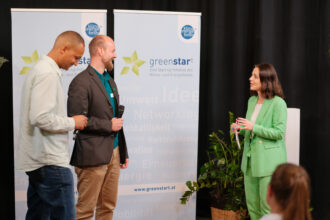 Drei Personen stehen auf einer Bühne vor einem „greenstart“-Banner, das eine nachhaltige Start-up-Initiative bewirbt. Eine Frau im grünen Anzug hält ein Dokument und spricht mit zwei Männern, von denen einer ein Mikrofon in der Hand hält. Die Szene wirkt wie eine Preisverleihung oder eine Ehrung im Bereich Umwelt und Innovation.