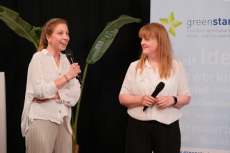 Zwei Frauen stehen auf einer Bühne vor einem „greenstart“-Banner, das eine nachhaltige Start-up-Initiative bewirbt. Beide halten Mikrofone in den Händen, während die Frau auf der linken Seite spricht und die andere aufmerksam zuhört. Die Szene wirkt wie eine Präsentation oder ein Gespräch über nachhaltige Innovationen.