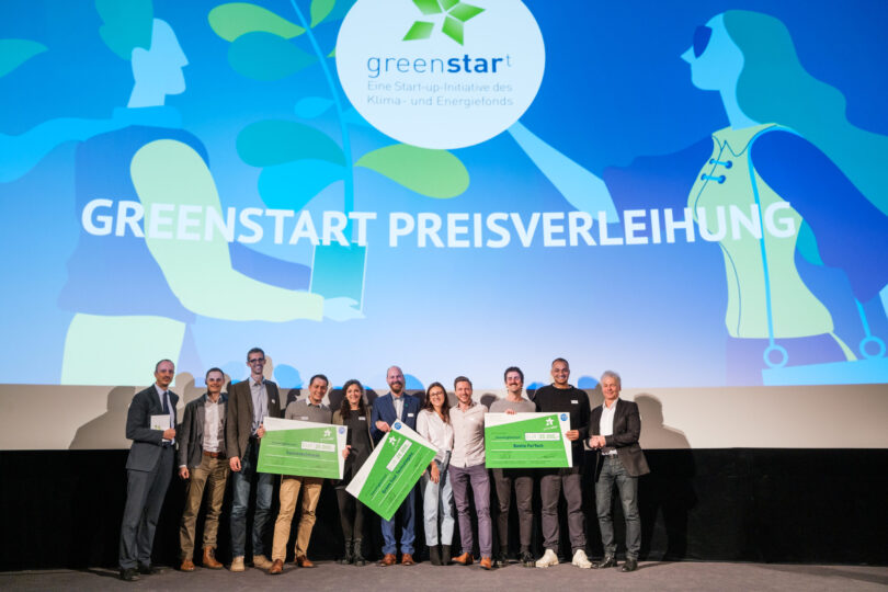 Eine Gruppe von Gewinnern bei der Greenstart-Preisverleihung, die große Schecks präsentieren. Im Hintergrund ein leuchtend blauer Bildschirm mit dem Greenstart-Logo und dem Text 'Greenstart Preisverleihung'.