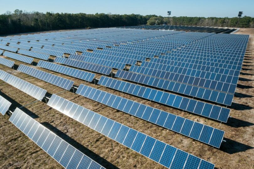 Ein großes Solarfeld mit hunderten von Solarmodulen, die in ordentlichen Reihen aufgestellt sind und das Sonnenlicht reflektieren. Das Feld liegt auf trockenem Grasland, umgeben von einem Wald im Hintergrund unter einem klaren blauen Himmel.
