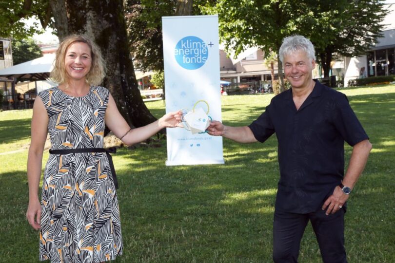 Eine Frau und ein Mann stehen auf einer grünen Wiese im Freien und lächeln in die Kamera. Sie halten gemeinsam ein weißes, herzförmiges Objekt mit einem gelben Griff. Im Hintergrund steht ein Banner mit dem Logo des 