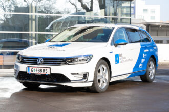 Ein weiß-blaues Auto, ein Volkswagen Passat GTE, ist von vorne links zu sehen. Auf der Motorhaube und den Türen befinden sich blaue Beschriftungen mit dem Logo von AVL und dem Text 'AVL KEYTECHAEV FUEL CELL DEMONSTRATOR'. Das Fahrzeug hat ein österreichisches Kennzeichen mit der Nummer 'G 468 RS'. Im Hintergrund ist ein modernes Gebäude mit Glasfassade zu erkennen, das sich in der glänzenden Oberfläche des Autos spiegelt.