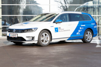Ein weiß-blaues Auto, ein Volkswagen Passat GTE, ist von vorne links zu sehen. Auf der Motorhaube und den Türen befinden sich blaue Beschriftungen mit dem Logo von AVL und dem Text 'AVL KEYTECHAEV FUEL CELL DEMONSTRATOR'. Das Fahrzeug hat ein österreichisches Kennzeichen mit der Nummer 'G 468 RS'. Im Hintergrund ist ein modernes Gebäude mit Glasfassade zu erkennen, das sich in der glänzenden Oberfläche des Autos spiegelt.
