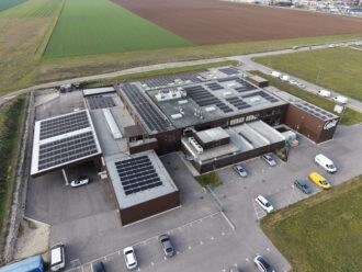 Luftbildaufnahme eines Produktionsstandortes, auf dessen Dächer PV Module montiert sind und auf dem Parkplatz mehrere Fahrzeuge stehen.