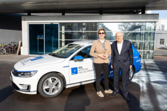 Zwei Personen stehen vor einem weißen Auto mit blauer Beschriftung, das als 'AVL KEYTECHAEV FUEL CELL DEMONSTRATOR' gekennzeichnet ist. Die Frau trägt eine beige Jacke und eine Sonnenbrille, der Mann einen dunklen Anzug. Im Hintergrund befindet sich ein modernes Gebäude mit Glasfassade, auf der das Logo von AVL sichtbar ist. Links sind einige Fahrräder abgestellt.