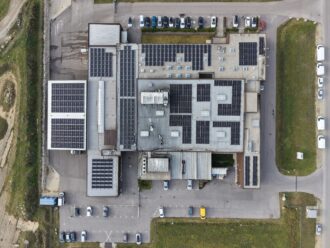 Luftbildaufnahme eines Produktionsstandortes, auf dessen Dächer PV Module montiert sind und auf dem Parkplatz mehrere Fahrzeuge stehen.