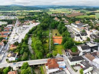 Luftaufnahme einer Kleinstadt, die von grünen Feldern und sanften Hügeln umgeben ist. In der Bildmitte verläuft eine Baumallee, die an Tennisplätzen und einer Kirche mit gelber Fassade vorbeiführt. Links davon schlängelt sich ein Fluss durch die Stadt, begleitet von einem Parkplatz und Wohngebäuden. Im Hintergrund erstreckt sich die ländliche Landschaft mit Feldern, Wiesen und verstreuten Häusern unter einem bewölkten Himmel.