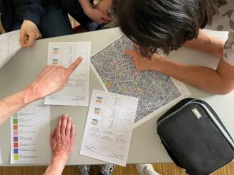 Mehrere Personen sitzen an einem Tisch und analysieren eine Stadtkarte, auf der verschiedene Bereiche farblich markiert sind. Sie zeigen mit den Fingern auf bestimmte Punkte der Karte, während auf dem Tisch auch Arbeitsblätter mit Diagrammen und einer Farblegende liegen. Eine schwarze Tasche mit der Aufschrift „TUSENT“ liegt ebenfalls auf dem Tisch. Die Szene deutet auf eine gemeinsame Planungs- oder Rechercheaufgabe hin.