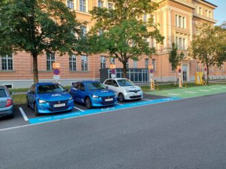 Drei Parkende E-Autos an Ladestationen. Im Hintergrund Bäume, ein Energiespeicher und ein Gebäude.