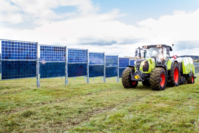 Ein Traktor neben einer Reihe von Solarmodulen auf einer Wiese unter einem bewölkten Himmel.