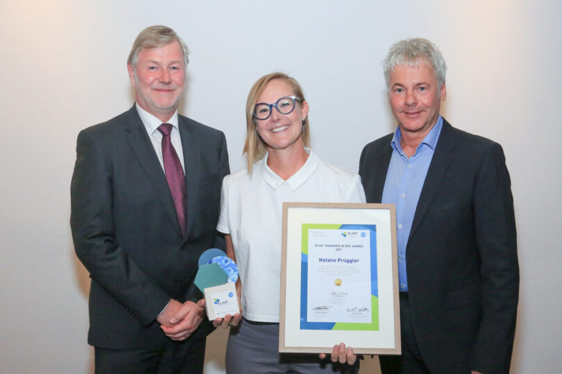 Drei Personen posieren für ein Foto bei einer Preisverleihung. In der Mitte hält eine Frau mit Brille ein gerahmtes Zertifikat mit der Aufschrift „KLAR!-Managerin des Jahres 2023“ und dem Namen „Natalie Prügger“. In ihrer anderen Hand hält sie eine Trophäe mit einem blauen und grünen Design. Die beiden Männer neben ihr tragen Anzüge und lächeln in die Kamera. Der Hintergrund ist schlicht und neutral, um den Fokus auf die Personen zu lenken.