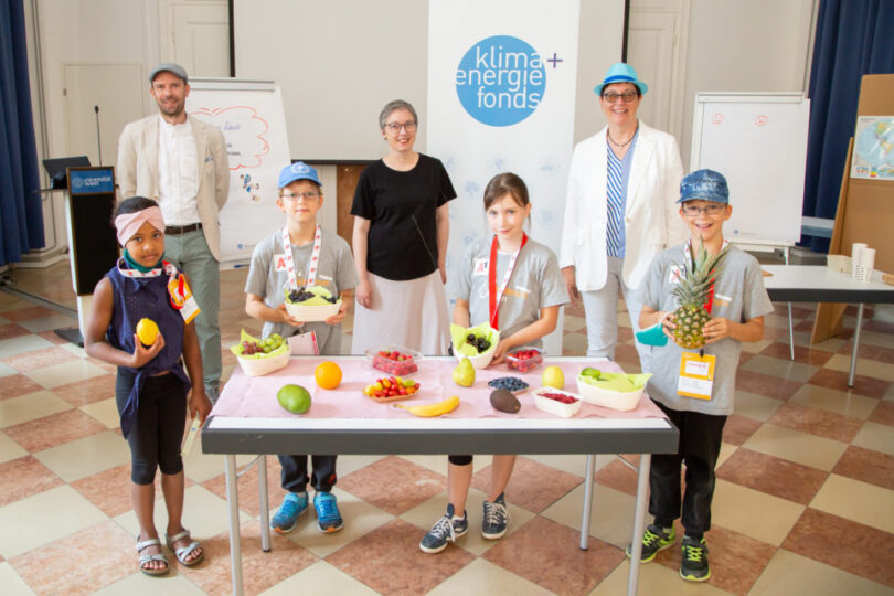 Eine Gruppe von fünf Kindern und drei Erwachsenen steht in einem Raum mit kariertem Boden. Die Kinder halten verschiedene Obstsorten wie Mango, Ananas, Beeren und Bananen in den Händen, während weiteres Obst auf einem Tisch vor ihnen liegt. Im Hintergrund befindet sich ein Roll-up-Banner mit dem Logo des 