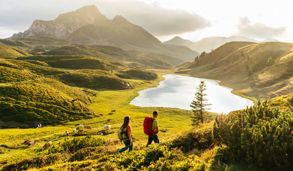 Zwei Wanderer mit Rucksäcken genießen eine malerische Landschaft in den Bergen, während sie einen grünen Hang entlanglaufen. Im Hintergrund erstrecken sich sanfte, grüne Hügel und ein klarer, schimmernder Bergsee, der von der untergehenden Sonne beleuchtet wird. Kühe grasen friedlich auf den saftigen Wiesen, umgeben von vereinzelten Bäumen und einer majestätischen Bergkulisse unter teils bewölktem Himmel.