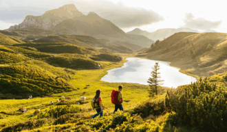 Zwei Wanderer mit Rucksäcken genießen eine malerische Landschaft in den Bergen, während sie einen grünen Hang entlanglaufen. Im Hintergrund erstrecken sich sanfte, grüne Hügel und ein klarer, schimmernder Bergsee, der von der untergehenden Sonne beleuchtet wird. Kühe grasen friedlich auf den saftigen Wiesen, umgeben von vereinzelten Bäumen und einer majestätischen Bergkulisse unter teils bewölktem Himmel.
