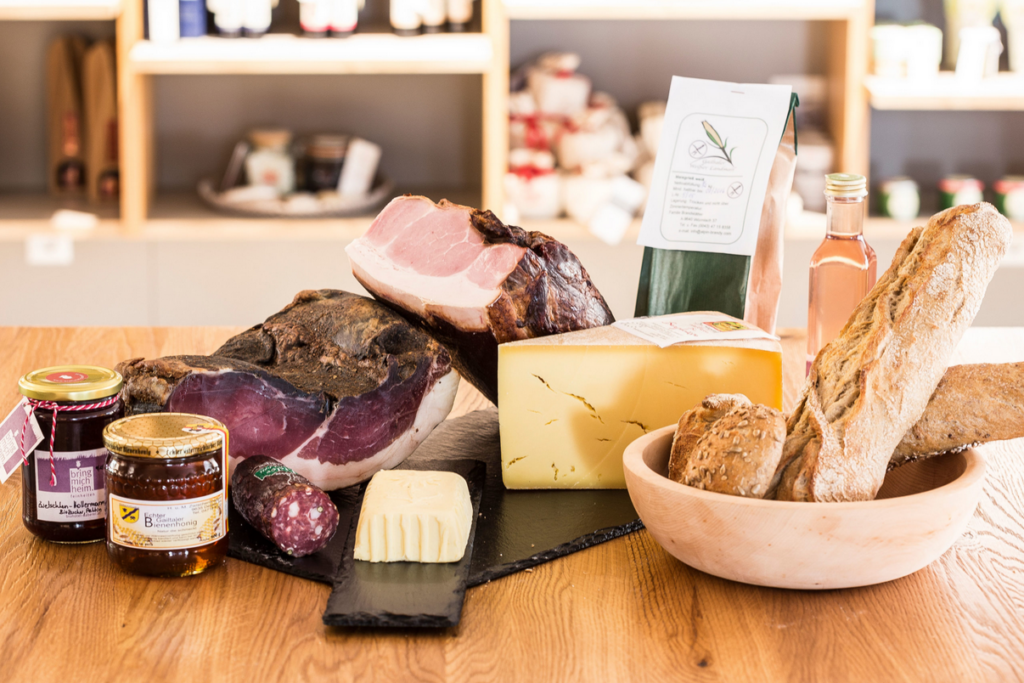 Ein rustikales Holztablett präsentiert eine Auswahl regionaler Delikatessen, darunter geräucherter Schinken, ein großes Stück Käse, eine kleine Salami, Butter, frisches Bauernbrot und Baguette. Ergänzt wird das Arrangement durch ein Glas Honig, ein Glas Marmelade, eine Flasche Roséwein und eine Packung Kräutertee. Im Hintergrund sind Regale mit weiteren regionalen Produkten unscharf zu erkennen.