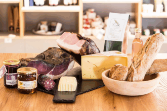 Ein rustikales Holztablett präsentiert eine Auswahl regionaler Delikatessen, darunter geräucherter Schinken, ein großes Stück Käse, eine kleine Salami, Butter, frisches Bauernbrot und Baguette. Ergänzt wird das Arrangement durch ein Glas Honig, ein Glas Marmelade, eine Flasche Roséwein und eine Packung Kräutertee. Im Hintergrund sind Regale mit weiteren regionalen Produkten unscharf zu erkennen.