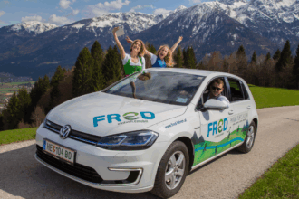 Ein weißes Elektroauto der Marke Volkswagen mit der Aufschrift 'FRED einfach.emobil' steht auf einer schmalen Landstraße inmitten einer idyllischen Berglandschaft. Im Hintergrund ragen schneebedeckte Alpen majestätisch in den Himmel, flankiert von dichten, grünen Tannenwäldern. Zwei fröhliche junge Frauen lehnen sich lachend aus dem Schiebedach des Autos, eine von ihnen hält ein Smartphone für ein Selfie. Ein junger Mann sitzt lässig am Steuer und lächelt in die Kamera. Die Szene strahlt Lebensfreude, Nachhaltigkeit und Abenteuerlust aus.