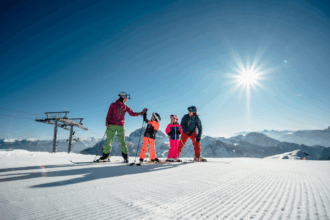 Eine Familie steht in bunter Skibekleidung auf einer perfekt präparierten Piste, umgeben von einer beeindruckenden Alpenlandschaft unter strahlend blauem Himmel. Die Sonne scheint hell und wirft lange Schatten auf den Schnee. Zwei Kinder in leuchtend orangener und pinker Skiausrüstung stehen zwischen den Eltern, die liebevoll mit ihnen interagieren. Im Hintergrund sind schneebedeckte Berggipfel und ein Skilift zu sehen, der sich über das Panorama erstreckt. Die Szene vermittelt Lebensfreude, Familienzusammenhalt und Winterspaß in einer malerischen Bergwelt.