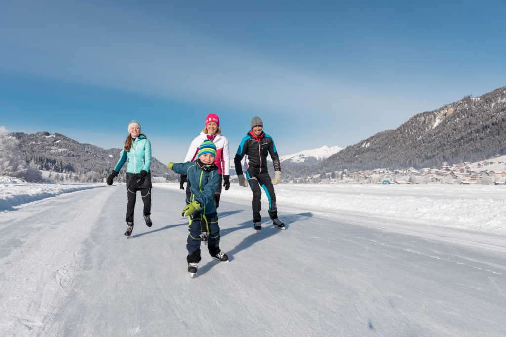 Eine fröhliche Familie aus vier Personen läuft Schlittschuh auf einer zugefrorenen Eisfläche inmitten einer verschneiten Winterlandschaft. Im Vordergrund ist ein lachender Junge in blauer Winterkleidung und bunter Mütze zu sehen, der freudig in die Kamera blickt. Hinter ihm laufen zwei Erwachsene und ein weiteres Kind, alle warm angezogen. Im Hintergrund sind schneebedeckte Berge, Tannenwälder und ein sonniger, blauer Himmel zu erkennen.