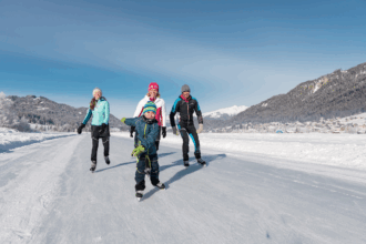 Eine fröhliche Familie aus vier Personen läuft Schlittschuh auf einer zugefrorenen Eisfläche inmitten einer verschneiten Winterlandschaft. Im Vordergrund ist ein lachender Junge in blauer Winterkleidung und bunter Mütze zu sehen, der freudig in die Kamera blickt. Hinter ihm laufen zwei Erwachsene und ein weiteres Kind, alle warm angezogen. Im Hintergrund sind schneebedeckte Berge, Tannenwälder und ein sonniger, blauer Himmel zu erkennen.
