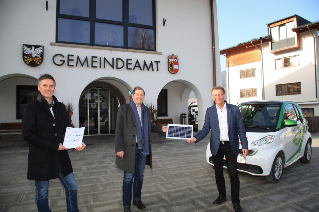 Drei Männer stehen vor dem Gebäude eines Gemeindeamts mit den Wappen von Österreich und einem Bundesland. Der Mann links hält ein Dokument, der mittlere Mann lächelt in die Kamera, während der rechte Mann ein kleines Solarpanel präsentiert. Im Hintergrund steht ein kleines Elektroauto mit grüner Beschriftung, das auf Nachhaltigkeit und umweltfreundliche Mobilität hinweist. Das Bild vermittelt eine Szene im Zusammenhang mit erneuerbarer Energie und umweltbewusster Kommunalarbeit.