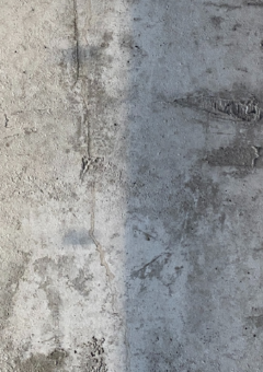 Detailaufnahme einer grauen Betonwand mit sichtbaren Rissen, Kratzern und leichten Verschmutzungen. Die Oberfläche zeigt eine Mischung aus rauen und glatten Texturen sowie verschiedene Schattierungen von Grau.