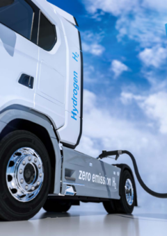 Ein weißer Wasserstoff-LKW mit der Aufschrift 'zero emission H₂' wird an einer Wasserstoff-Tankstelle betankt. Die Zapfsäule ist blau-weiß und mit 'H₂ Hydrogen' beschriftet, vor einem sonnigen Himmel mit weißen Wolken.