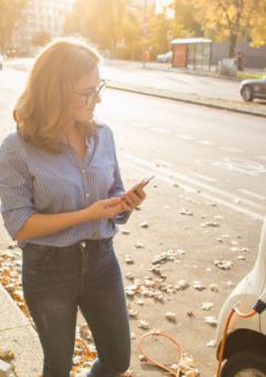 Eine Frau steht neben einem weißen Elektroauto, das an einer Ladestation aufgeladen wird. Sie schaut auf ihr Smartphone, während im Hintergrund eine sonnige Stadtstraße mit herbstlichem Laub zu sehen ist.