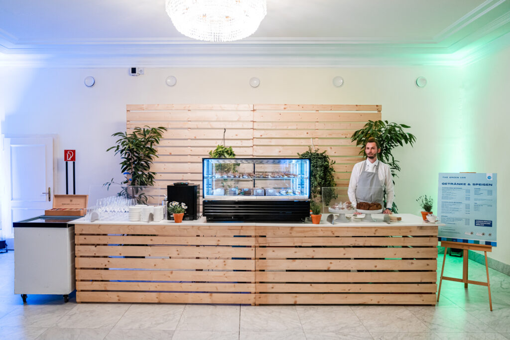 Das Bild zeigt eine nachhaltig gestaltete Catering-Station mit einer Theke aus hellem Holz und dekorativen Pflanzen. In der Mitte steht eine gekühlte Vitrine, die verschiedene Speisen präsentiert, darunter Sandwiches und Desserts. Rechts daneben befindet sich ein Barista oder Caterer in einer weißen Schürze, der hinter der Theke steht. Auf der rechten Seite ist eine Speisekarte mit dem Titel 