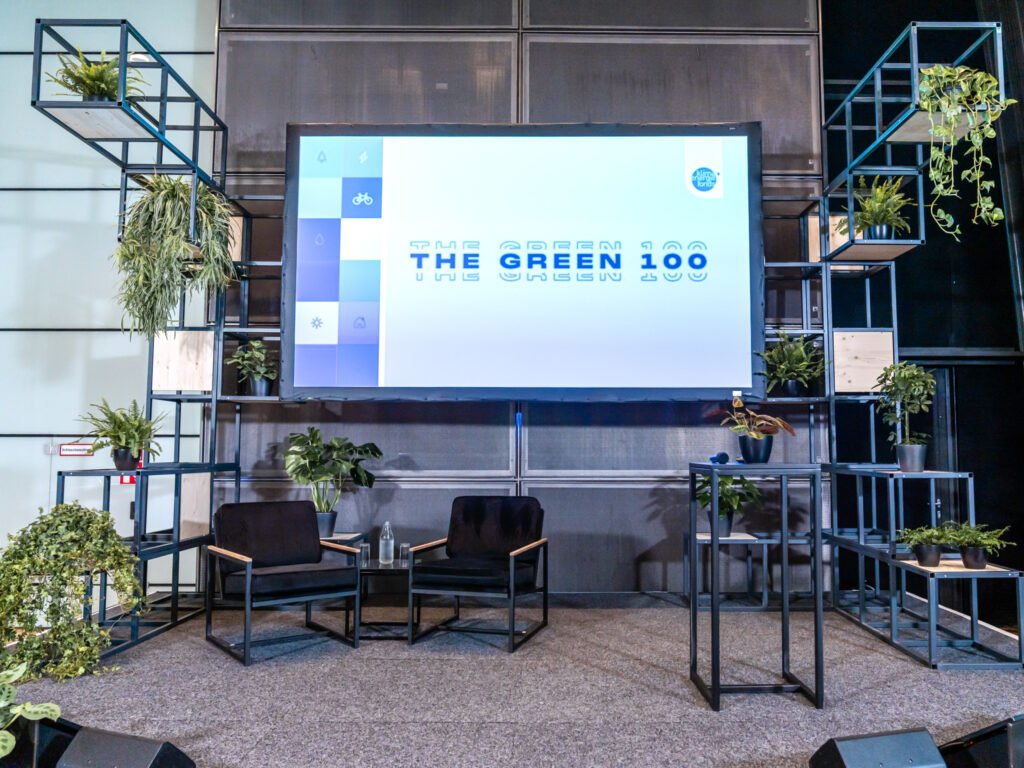 Das Bild zeigt eine Bühne für eine Veranstaltung mit dem Titel The Green 100, projiziert auf eine große Leinwand. Die Bühne ist modern gestaltet und mit Pflanzen dekoriert, die in schwarzen Metallregalen angeordnet sind, was eine nachhaltige und natürliche Ästhetik vermittelt. Im Vordergrund stehen zwei schwarze Sessel mit Holzarmlehnen sowie ein kleiner Tisch mit einer Pflanze. Die Bühne ist mit grauem Teppich ausgelegt, und die dunkle Metallwand im Hintergrund sorgt für einen modernen, industriellen Look. Das Bühnen-Setup wirkt einladend und professionell, passend für eine Podiumsdiskussion oder eine Präsentation zum Thema Nachhaltigkeit und grüne Innovationen.