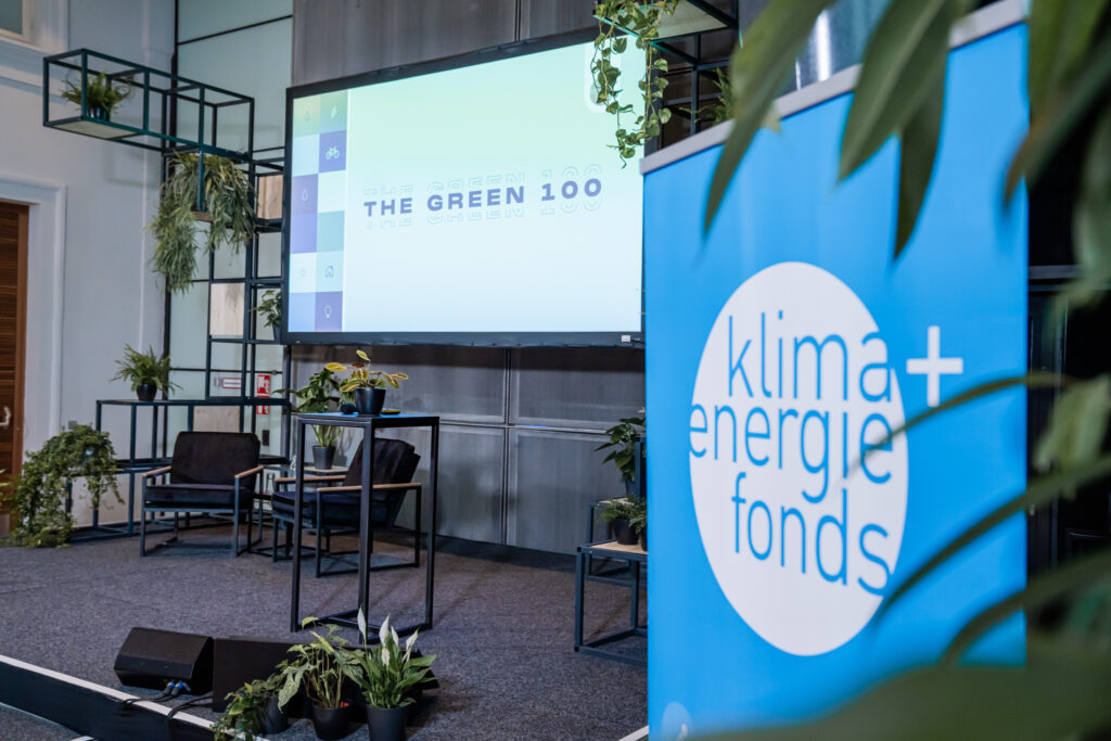 Das Bild zeigt eine Bühne für eine Präsentation oder Podiumsdiskussion mit dem Thema The Green 100. Im Vordergrund steht ein blaues Banner mit dem Logo von 