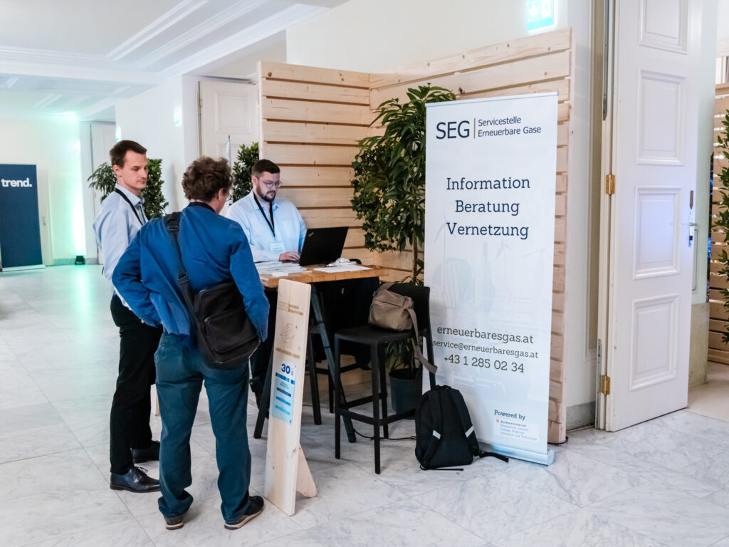 Das Bild zeigt den Messestand der Servicestelle Erneuerbare Gase (SEG). Ein großes weiß-blaues Banner informiert über die Dienstleistungen der SEG, darunter Information, Beratung und Vernetzung. Die Website erneuerbaresgas.at sowie eine E-Mail-Adresse und Telefonnummer sind darauf angegeben. Am Tisch stehen drei Männer in Business-Kleidung. Einer von ihnen sitzt am Laptop und scheint eine Beratung durchzuführen, während die anderen beiden interessiert zuhören. Ein schwarzer Rucksack und eine Umhängetasche liegen auf einem Stuhl neben dem Tisch. Die Standnummer 30 ist auf einer hölzernen Stele erkennbar, und die Rückwand besteht aus Holzpaneelen mit grünen Pflanzen, was eine natürliche und nachhaltige Atmosphäre vermittelt. Im Hintergrund sind weitere Messestände zu sehen, was darauf hinweist, dass sich die Veranstaltung auf erneuerbare Energien und nachhaltige Technologien konzentriert.
