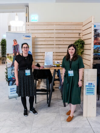 Das Bild zeigt den Messestand von Oikocredit, einer Organisation, die sich für soziale Investitionen und Armutsbekämpfung einsetzt. Zwei Frauen mit Namensschildern und dunklen Kleidern stehen vor dem Stand, lächeln in die Kamera und präsentieren das Informationsmaterial auf dem Holztisch. Hinter ihnen befindet sich ein großes Banner, das eine Frau in traditioneller Kleidung zeigt, mit dem Text „Geld für die Armutsbekämpfung“ und der Website www.oikocredit.at. Der Stand ist schlicht mit Holzwänden und grünen Pflanzen dekoriert, passend zum nachhaltigen und sozialen Engagement von Oikocredit. Auf dem Tisch liegt ein Ständer mit Flyern und Broschüren, die über die Projekte und Investitionsmöglichkeiten informieren. Eine Holzstele mit der Nummer 28 kennzeichnet den Stand von Oikocredit, und eine beige Stofftasche mit dem Logo der Organisation hängt an einem der Stühle. Das Gesamtbild vermittelt eine professionelle und einladende Atmosphäre mit dem Fokus auf ethische Finanzierung und soziale Verantwortung.