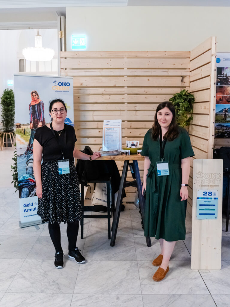 Das Bild zeigt den Messestand von Oikocredit, einer Organisation, die sich für soziale Investitionen und Armutsbekämpfung einsetzt. Zwei Frauen mit Namensschildern und dunklen Kleidern stehen vor dem Stand, lächeln in die Kamera und präsentieren das Informationsmaterial auf dem Holztisch. Hinter ihnen befindet sich ein großes Banner, das eine Frau in traditioneller Kleidung zeigt, mit dem Text „Geld für die Armutsbekämpfung“ und der Website www.oikocredit.at. Der Stand ist schlicht mit Holzwänden und grünen Pflanzen dekoriert, passend zum nachhaltigen und sozialen Engagement von Oikocredit. Auf dem Tisch liegt ein Ständer mit Flyern und Broschüren, die über die Projekte und Investitionsmöglichkeiten informieren. Eine Holzstele mit der Nummer 28 kennzeichnet den Stand von Oikocredit, und eine beige Stofftasche mit dem Logo der Organisation hängt an einem der Stühle. Das Gesamtbild vermittelt eine professionelle und einladende Atmosphäre mit dem Fokus auf ethische Finanzierung und soziale Verantwortung.