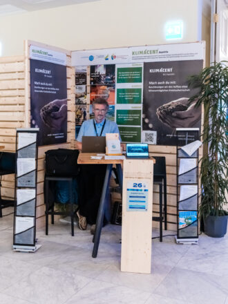 Das Bild zeigt den Messestand von KLIMACENT Austria, einer Initiative zur Förderung einer klimaneutralen Kreislaufwirtschaft. Ein Mann mit Brille und blauem Hemd sitzt hinter einem Stehtisch, arbeitet an seinem Laptop und lächelt in die Kamera. Der Stand ist mit zwei großen Bannern versehen, die den Slogan „Wir handeln“ tragen und zur Unterstützung des Projekts aufrufen. Auf dem Tisch stehen ein kleiner Laptop und eine Infotafel mit einem QR-Code, vermutlich zum Abrufen weiterer Informationen. Eine Holzstele mit der Nummer 26 kennzeichnet den Stand von KLIMACENT. Links und rechts vom Tisch befinden sich zwei schwarze Prospektständer, die verschiedene Flyer und Informationsblätter über das Projekt enthalten. Die Gestaltung des Standes nutzt natürliche Materialien wie Holz und Pflanzen, was den nachhaltigen Fokus der Initiative unterstreicht. Das Ambiente wirkt professionell und einladend, mit gut sichtbaren Informationstafeln zur Darstellung der Klimaziele und Maßnahmen von KLIMACENT.