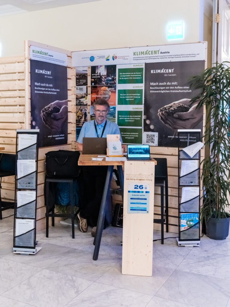 Das Bild zeigt den Messestand von KLIMACENT Austria, einer Initiative zur Förderung einer klimaneutralen Kreislaufwirtschaft. Ein Mann mit Brille und blauem Hemd sitzt hinter einem Stehtisch, arbeitet an seinem Laptop und lächelt in die Kamera. Der Stand ist mit zwei großen Bannern versehen, die den Slogan „Wir handeln“ tragen und zur Unterstützung des Projekts aufrufen. Auf dem Tisch stehen ein kleiner Laptop und eine Infotafel mit einem QR-Code, vermutlich zum Abrufen weiterer Informationen. Eine Holzstele mit der Nummer 26 kennzeichnet den Stand von KLIMACENT. Links und rechts vom Tisch befinden sich zwei schwarze Prospektständer, die verschiedene Flyer und Informationsblätter über das Projekt enthalten. Die Gestaltung des Standes nutzt natürliche Materialien wie Holz und Pflanzen, was den nachhaltigen Fokus der Initiative unterstreicht. Das Ambiente wirkt professionell und einladend, mit gut sichtbaren Informationstafeln zur Darstellung der Klimaziele und Maßnahmen von KLIMACENT.