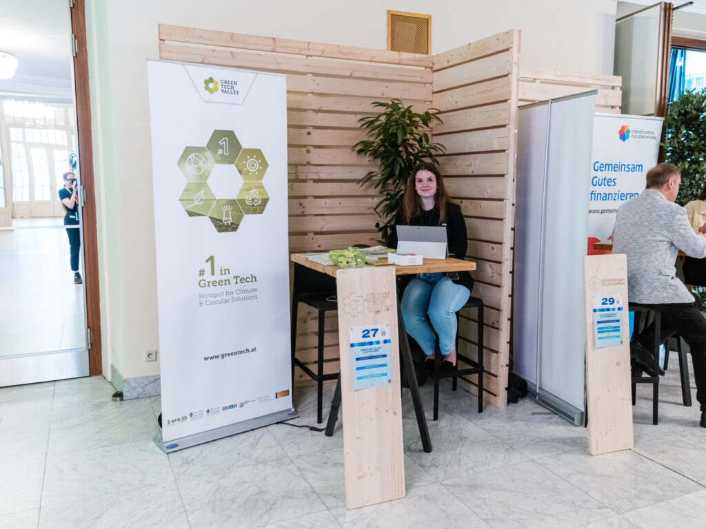 Das Bild zeigt den Messestand von Green Tech Valley, einem Hotspot für Klima- und Kreislauflösungen. Eine junge Frau sitzt an einem hohen Stehtisch, lächelt in die Kamera und hat einen Laptop vor sich. Auf dem Tisch liegen einige Flyer und Giveaways, die vermutlich über nachhaltige Technologien und Innovationen informieren. Links neben ihr steht ein großes Banner mit sechseckigen Symbolen, das die verschiedenen Schwerpunkte von Green Tech Valley darstellt. Der Slogan „#1 in Green Tech – Hotspot for Climate & Circular Solutions“ hebt die zentrale Rolle dieser Initiative im Bereich Umwelttechnologie hervor. Die Website www.greentech.at ist ebenfalls auf dem Banner zu sehen. Rechts im Bild ist der Stand der Genossenschaft für Gemeinwohl mit dem Slogan „Gemeinsam Gutes finanzieren“ teilweise sichtbar. Beide Stände sind durch Holztrennwände und grüne Pflanzen umgeben, was eine natürliche und nachhaltige Atmosphäre schafft. Eine Holzstele mit der Nummer 27 kennzeichnet den Stand von Green Tech Valley.