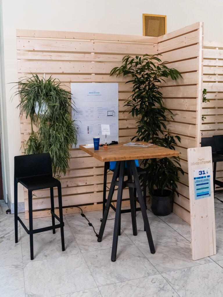 Das Bild zeigt einen Messestand mit der Nummer 31, der aktuell unbesetzt ist. Der Stand besteht aus einem hohen Stehtisch mit zwei schwarzen Barhockern und ist von einer hölzernen Rückwand mit grünen Pflanzen umgeben, was eine natürliche Atmosphäre schafft. An der Rückwand hängt ein Papier mit einer Skizze oder einem Informationsplan, vermutlich mit Details zu einem Projekt oder einer Initiative. Auf dem Tisch liegen eine blaue Tasse mit Logo, ein kleines Schild, einige Unterlagen und Stifte, die möglicherweise für Besucher oder Präsentationen gedacht sind. Die Holzstele rechts im Bild trägt den Namen des Ausstellers, allerdings ist der Schriftzug nicht vollständig lesbar. Insgesamt vermittelt der Stand einen minimalistischen und nachhaltigen Eindruck, passend zur natürlichen Gestaltung der Umgebung.