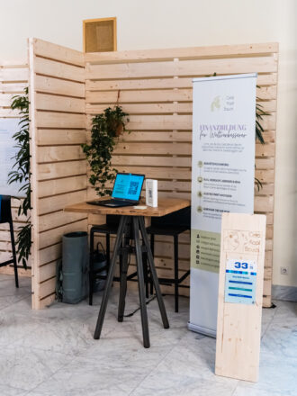 Das Bild zeigt den Messestand von „Geld Kopf Bauch“, einem Anbieter für Finanzbildung für Weltverbesserer. Ein großes, helles Banner hebt verschiedene Angebote hervor, darunter Investitionsberatung, Bildungsangebote wie Workshops und Webinare, selbstbestimmtes Investieren sowie gemeinschaftliche Finanzlösungen. In der Mitte des Standes steht ein hoher Stehtisch mit zwei schwarzen Barhockern. Auf dem Tisch befindet sich ein Laptop mit einer offenen Website sowie ein QR-Code für weitere Informationen. Das Holzpaneel im Hintergrund ist mit grünen Pflanzen dekoriert, was dem Stand eine natürliche, freundliche Atmosphäre verleiht. Rechts im Bild ist eine Holzstele mit der Nummer 33 zu sehen, die den Stand kennzeichnet. Unter dem Tisch steht eine Tasche oder Rollbehälter, was darauf hindeutet, dass Materialien oder persönliche Gegenstände verstaut wurden.