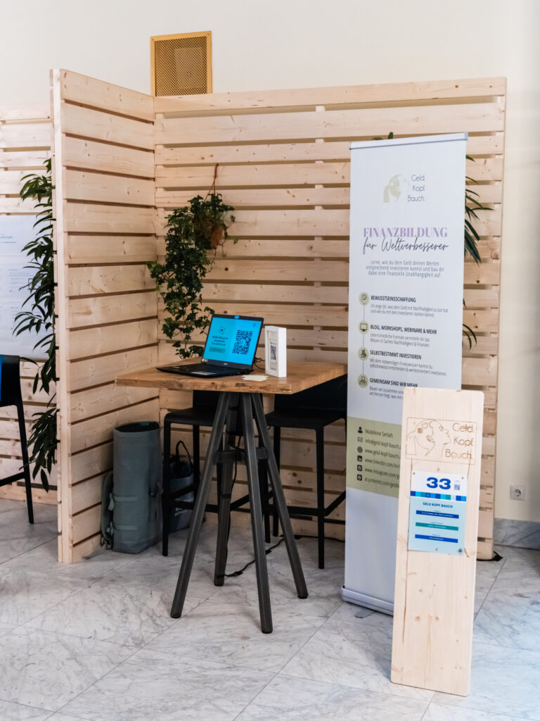 Das Bild zeigt den Messestand von „Geld Kopf Bauch“, einem Anbieter für Finanzbildung für Weltverbesserer. Ein großes, helles Banner hebt verschiedene Angebote hervor, darunter Investitionsberatung, Bildungsangebote wie Workshops und Webinare, selbstbestimmtes Investieren sowie gemeinschaftliche Finanzlösungen. In der Mitte des Standes steht ein hoher Stehtisch mit zwei schwarzen Barhockern. Auf dem Tisch befindet sich ein Laptop mit einer offenen Website sowie ein QR-Code für weitere Informationen. Das Holzpaneel im Hintergrund ist mit grünen Pflanzen dekoriert, was dem Stand eine natürliche, freundliche Atmosphäre verleiht. Rechts im Bild ist eine Holzstele mit der Nummer 33 zu sehen, die den Stand kennzeichnet. Unter dem Tisch steht eine Tasche oder Rollbehälter, was darauf hindeutet, dass Materialien oder persönliche Gegenstände verstaut wurden.